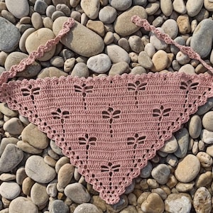 Può includere: Una bandana all'uncinetto rosa polvere con un motivo a libellula e bordi smerlati. La bandana ha due lunghi lacci per fissarla intorno al collo o alla testa. L'accessorio fatto a mano è esposto su un letto di pietre lisce.