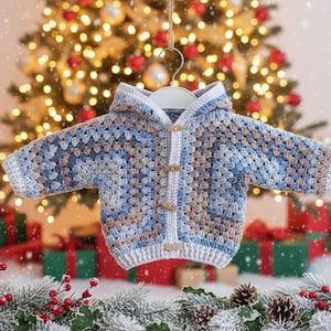 May include: A hooded cardigan featuring a crochet granny square pattern in shades of blue, white, and tan. The garment includes a white crocheted border along the front opening, long sleeves, and four light brown wooden toggle buttons for closure.