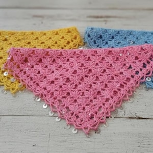 May include: Three handmade crochet bandanas in yellow, pink, and blue. Each bandana is triangular with a floral pattern and embellished with sequins along the edges. The bandanas are displayed on a weathered, white wooden surface.