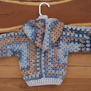 May include: A hooded sweater crafted with a crochet granny square pattern in shades of light blue, dark blue, and tan. The garment features long sleeves with ribbed cuffs, a ribbed hem, and a hood, all constructed from yarn in a multi-colored design.