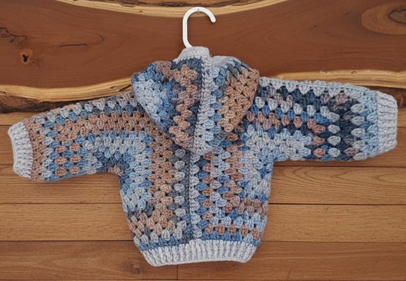 May include: A hooded sweater crafted with a crochet granny square pattern in shades of light blue, dark blue, and tan. The garment features long sleeves with ribbed cuffs, a ribbed hem, and a hood, all constructed from yarn in a multi-colored design.