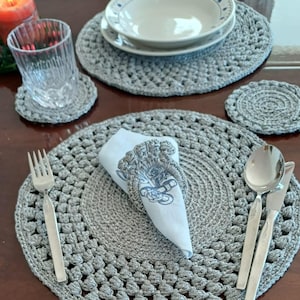 May include: A dining table setting featuring crocheted gray placemats, coasters, and a napkin ring. The setting includes a bowl, plate, glass, silverware, and a folded napkin. A lit candle is visible in the background.