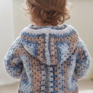 May include: A hooded crochet sweater featuring a multi-colored granny square pattern in shades of dusty blue, light grey, and muted brown. The garment has long sleeves, a hood, and a textured yarn construction with a visible stitch pattern throughout.