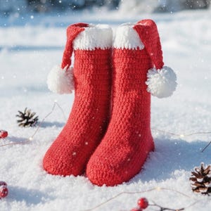 Puede incluir: Dos calcetines de Navidad rojos de ganchillo con ribete blanco y pompones, que se asemejan a gorros de Papá Noel, están en la nieve. Piñas y bayas rojas están esparcidas alrededor, creando una escena invernal festiva.