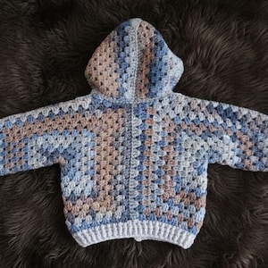 May include: A long-sleeve hooded sweater crafted with a granny square crochet pattern. The garment features a color palette of muted blue, light grey, and soft tan tones, finished with a solid light grey ribbed hem and sleeve cuffs.