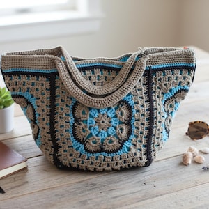 May include: A handmade crochet tote bag in shades of tan, black, and light blue. The bag features a floral design on the front and a sturdy handle. A small succulent in a white pot, a brown notebook, and sunglasses are next to the bag.