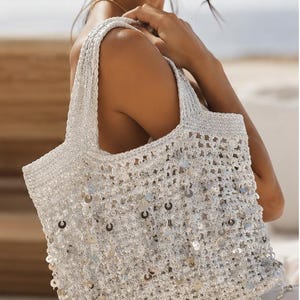 May include: A white crocheted tote bag featuring an open-weave construction adorned with numerous small, reflective silver sequins throughout the body and a fringe of dangling sequins along the bottom edge, complete with two sturdy crocheted handles.