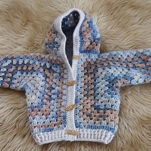 Könnte beinhalten: Gehäkelter Baby-Cardigan mit Kapuze im Granny-Square-Muster. Die Jacke hat Blau-, Beige- und Weißtöne und wird mit Holzknebelknöpfen geschlossen. Sie wird auf einer flauschigen, hellbraunen Oberfläche präsentiert.