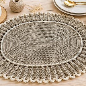Op de afbeelding: Een ovaal gehaakt placemat met een effen taupekleurig midden in een gestructureerd steekpatroon, omringd door een brede, geplooide taupekleurige rand en afgewerkt met een delicate, crèmekleurige schulprand.