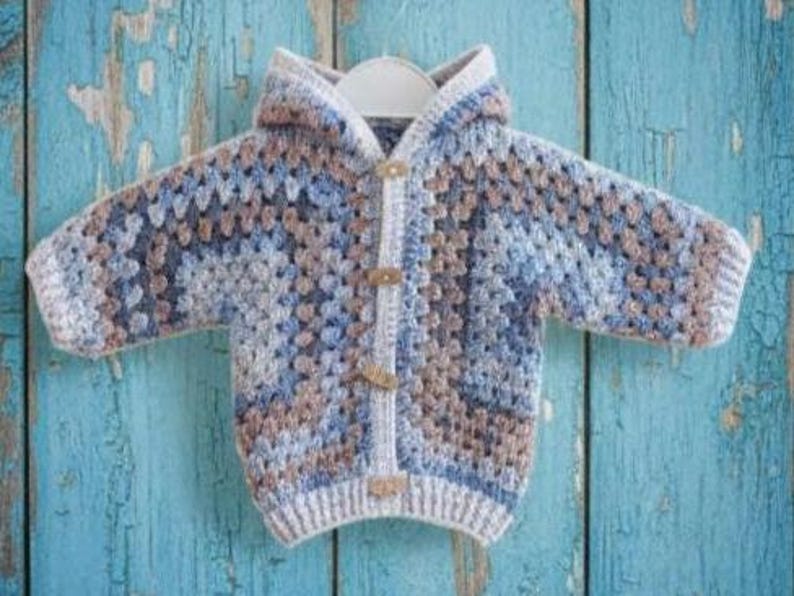 May include: A long-sleeve hooded cardigan featuring a multi-colored granny square crochet pattern in shades of blue, grey, and tan. The garment includes a front button closure with three light-colored wooden toggles and ribbed trim at the cuffs.