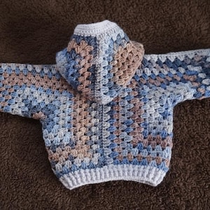 May include: A hooded sweater crafted with a crochet granny square pattern in shades of blue, tan, and white. The garment features long sleeves, a hood, and solid white ribbed trim along the cuffs, the bottom hem, and the edge of the hood opening.