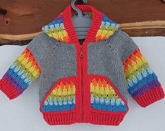Könnte beinhalten: Graue gehäkelte Kapuzenjacke mit rotem Reißverschluss und Besatz. Die Ärmel und Taschen weisen ein Regenbogenmuster aus Gelb, Orange, Blau und Grün auf. Die Jacke wird an einem grauen Kleiderbügel präsentiert.