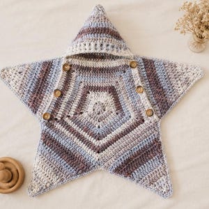 May include: A star-shaped crochet swaddle blanket featuring a hood and five wooden buttons. The textile displays a variegated pattern of cream, light blue, and muted brown yarn, constructed with a consistent stitch throughout the entire piece.