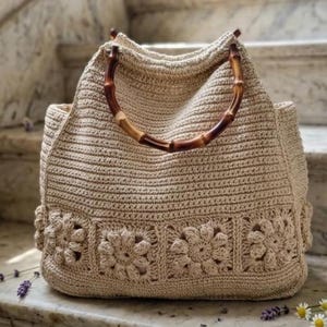 May include: A beige crochet tote bag with bamboo handles. The bag features a floral pattern along the bottom edge. The bag is sitting on a marble surface with lavender and daisies.