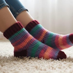 Puede incluir: Un par de calcetines tejidos a mano con un patrón de rayas en tonos morado, azul, verde, naranja y rosa. Los puños y los dedos son de color burdeos oscuro. Los calcetines están sobre una alfombra blanca.