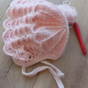 May include: A handmade, light pink crochet bonnet with a scalloped brim and white trim. The bonnet has tie strings and is next to a ball of matching yarn and a red crochet hook. The bonnet is likely for a baby.