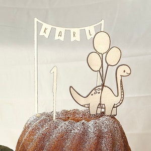 May include: A white cake stand with a white cake decorated with powdered sugar. A wooden dinosaur cake topper with balloons and the name "KARL" is on top of the cake.