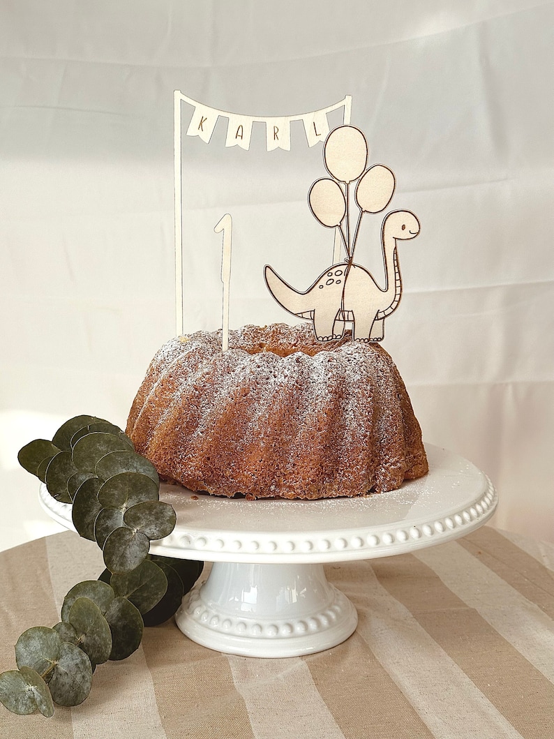 May include: A white cake stand with a white cake decorated with powdered sugar. A wooden dinosaur cake topper with balloons and the name "KARL" is on top of the cake.