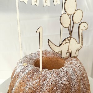 May include: Wooden cake topper featuring a dinosaur with balloons, a banner that says "KARL", and the number "1". The topper is on a cake with powdered sugar.