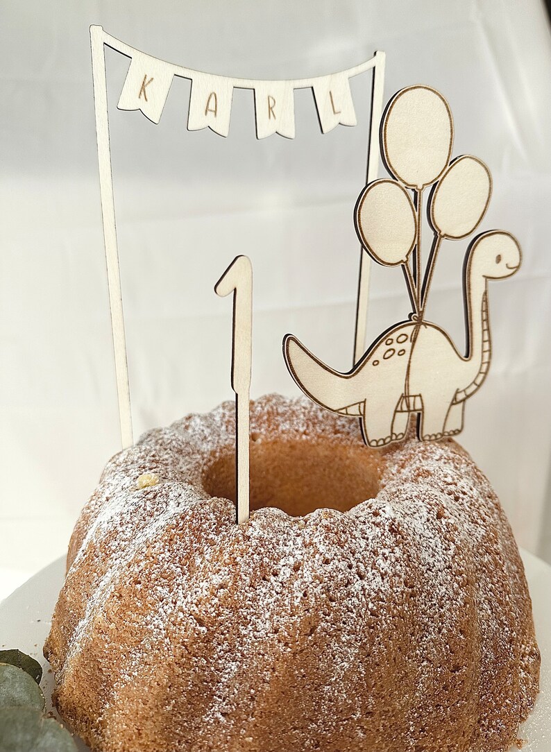 May include: Wooden cake topper featuring a dinosaur with balloons, a banner that says "KARL", and the number "1". The topper is on a cake with powdered sugar.