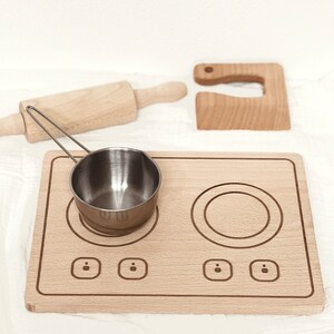 Könnte beinhalten: Holzspielzeug-Küchenset mit Herd, Nudelholz, kleinem Topf und Schneidebrett. Der Herd hat zwei Kochfelder und drei Knöpfe.