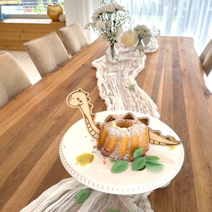 May include: A white cake decorated with green leaves and gold sprinkles sits on a white cake stand. A wooden dinosaur cake topper is placed on the cake. The cake stand is on a wooden table with a white tablecloth.
