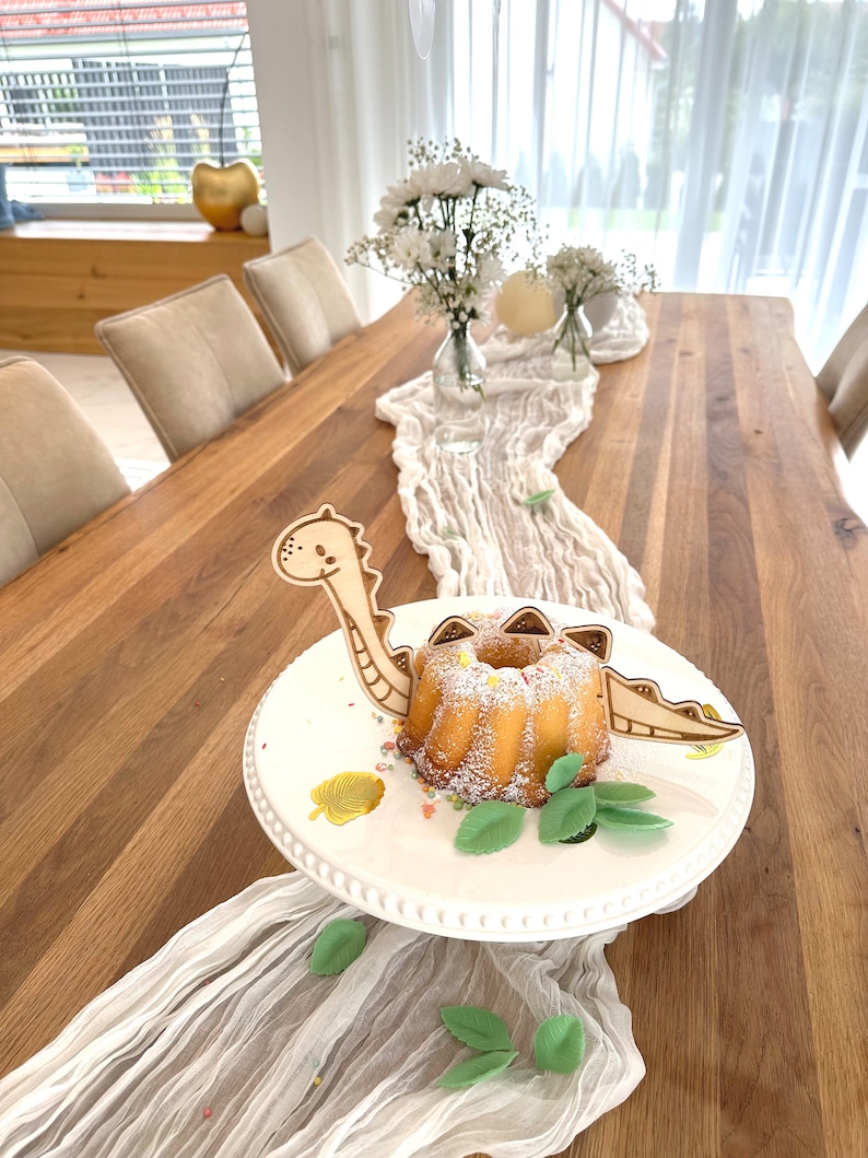 May include: A white cake decorated with green leaves and gold sprinkles sits on a white cake stand. A wooden dinosaur cake topper is placed on the cake. The cake stand is on a wooden table with a white tablecloth.