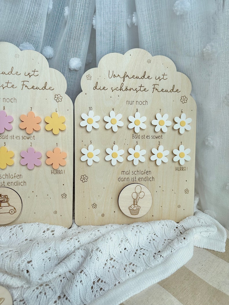 May include: Wooden countdown boards with daisy and flower cutouts. The boards have German text, including "Vorfreude ist die sch&ouml;nste Freude." One board has a car illustration, the other a cupcake with balloons.