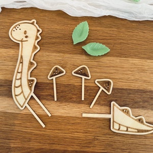 May include: Wooden craft kit featuring a dinosaur, mushrooms, and leaves. The pieces are cut out and ready to be assembled.