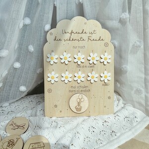 May include: Wooden countdown board with daisy-shaped markers and the German text "Vorfreude ist die sch&ouml;nste Freude." Includes small wooden tokens with illustrations. The board has a cloud-shaped top and is decorated with floral accents.