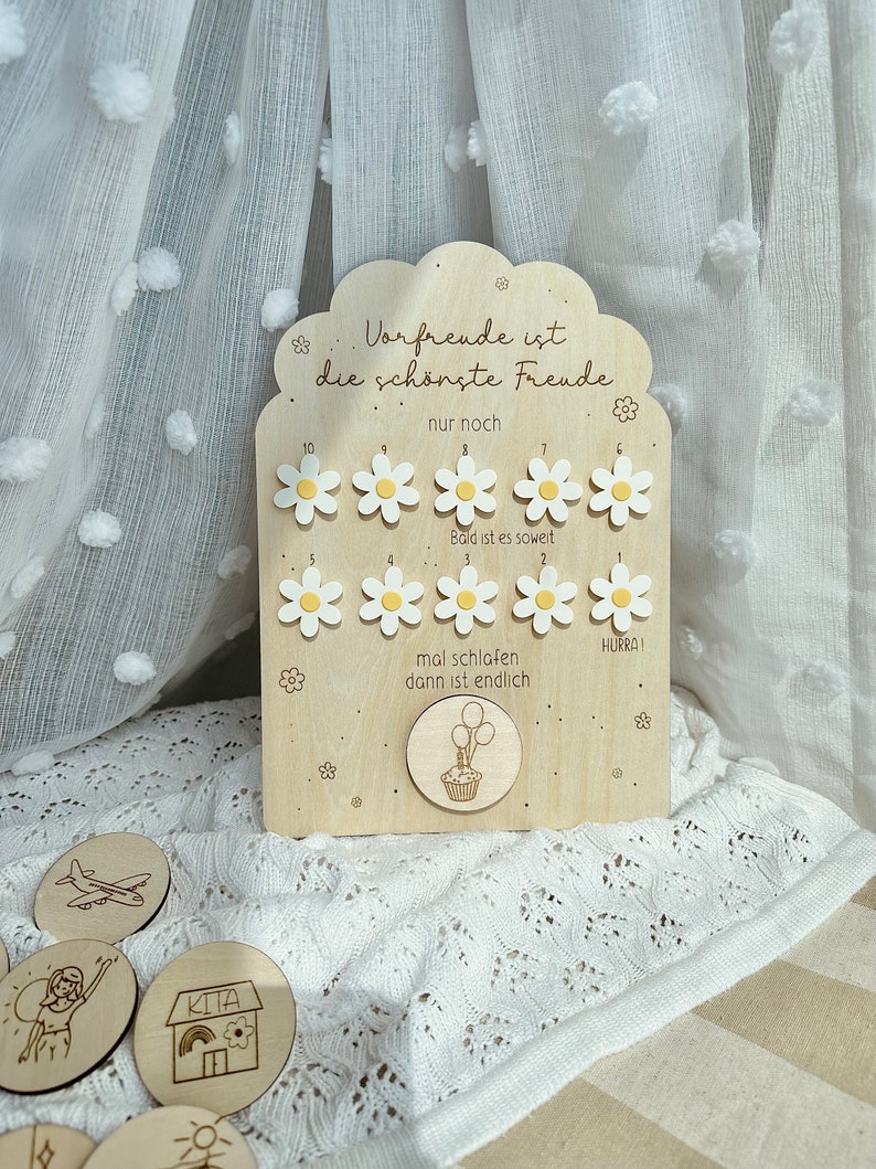 May include: Wooden countdown board with daisy-shaped markers and the German text "Vorfreude ist die sch&ouml;nste Freude." Includes small wooden tokens with illustrations. The board has a cloud-shaped top and is decorated with floral accents.