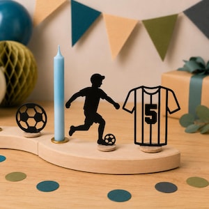 Könnte beinhalten: Ein heller Holzsockel trägt eine hellblaue Kerze, eine schwarze Silhouette eines Fußballs, eine schwarze Silhouette eines Fußballspielers beim Schuss sowie eine schwarze Silhouette eines Fußballtrikots mit der aufgedruckten Nummer 5.