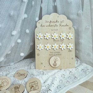 May include: Wooden countdown board with daisy-shaped markers and the text "Vorfreude ist die sch&ouml;nste Freude." Includes round wooden tokens with engraved illustrations. The board has a cupcake and balloon design.