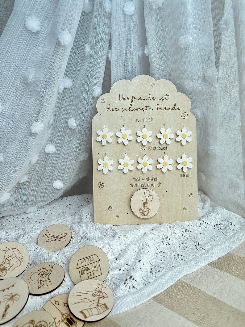 May include: Wooden countdown board with daisy-shaped markers and the text "Vorfreude ist die sch&ouml;nste Freude." Includes round wooden tokens with engraved illustrations. The board has a cupcake and balloon design.