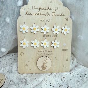 May include: Wooden countdown plaque with daisy accents. The plaque has the German text "Vorfrende ist die sch&ouml;nste Freude" and numbers 1-10 with daisy markers. A circular piece features a cupcake with balloons. A decorative item for a child.