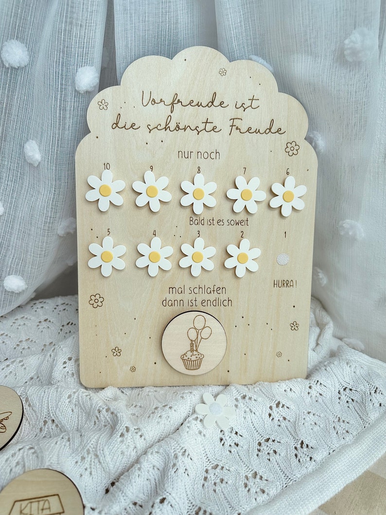 May include: Wooden countdown plaque with daisy accents. The plaque has the German text "Vorfrende ist die sch&ouml;nste Freude" and numbers 1-10 with daisy markers. A circular piece features a cupcake with balloons. A decorative item for a child.