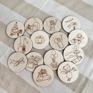 May include: A collection of fifteen wooden discs, each engraved with a different illustration. The designs include a Christmas tree, a bunny, a person waving, a palm tree, a jack-o'-lantern, a plane, a car with luggage, and a fairy.