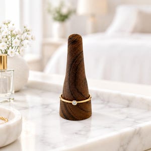 May include: A thin gold ring featuring a single small round clear gemstone set in a bezel, displayed on a dark brown wooden cone-shaped ring holder. The ring has a polished metallic finish and a simple, slender band design.