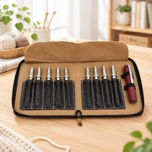 May include: A tan fabric zippered case containing eight silver metal crochet hooks with visible size markings and one dark red wooden handle. The hooks are organized in individual black textured slots inside the open, rectangular carrying case.