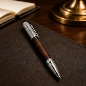 May include: A wooden pen with a silver tip and accents, resting on a brown leather surface. The pen has a dark wood barrel and a silver clip. The pen has the text "With God, Steadfast, Unshakeable, Thank You" engraved on the silver portion.