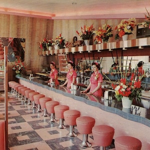 May include: A retro diner interior with a long counter and pink stools. Four women in pink dresses are standing behind the counter. The counter is decorated with a black and white checkered floor and a pink and white countertop. There are flowers on the shelves behind the counter.
