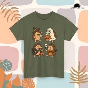 Op de afbeelding: Olijfgroen T-shirt met een Halloween-thema. De afbeelding toont vier cartoon egels verkleed als: een heks, een spook, een vampier en een mummie. Elke egel houdt een Halloween-prop vast, zoals een pompoen of een schedel.