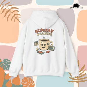 Op de afbeelding: Witte hoodie met een afbeelding op de achterkant. De afbeelding toont een verdrietige koffiekop met de woorden "SUNDAY SCARIES" erboven. Extra elementen zijn een kalender, een telefoon en een digitale klok.