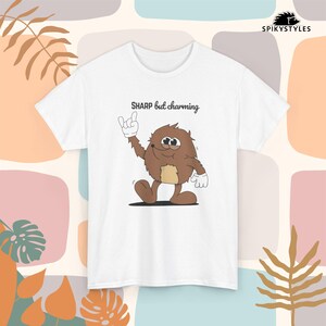 Op de afbeelding: Wit T-shirt met een cartoonfiguur met bruine vacht, een beige buik en witte handschoenen. De tekst "SHARP but charming" staat boven het personage. Het shirt heeft korte mouwen en een ronde hals.