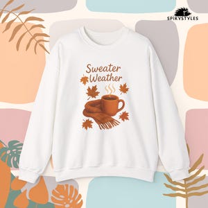 Op de afbeelding: Witte sweatshirt met de tekst "Sweater Weather", een koffiekopje, sjaal en herfstbladeren in bruin- en oranjetinten. Het ontwerp roept een gezellig herfstthema op.