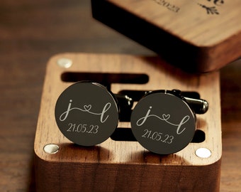 May include: A pair of black cufflinks with a silver inscription. The inscription reads "j&l 21.05.23". The cufflinks are in a wooden box.
