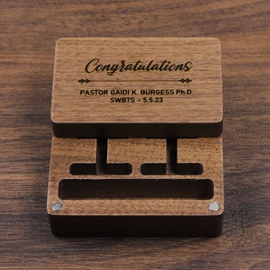 Può includere: Una scatola di legno con un coperchio incernierato. Il coperchio ha inciso il testo "Congratulations" e "PASTOR GAIDI K. BURGESS Ph.D. SWBTS - 5.5.23". La scatola ha tre scomparti.