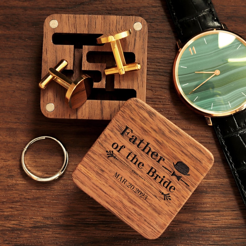 May include: A wooden box with a lid engraved with the text "Father of the Bride" and the date "MAR. 20. 2023". The box contains a pair of gold cufflinks and a silver ring.
