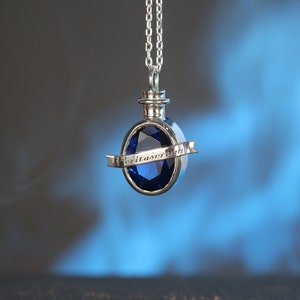 Puede incluir: Un collar de plata con un colgante de piedra azul. El colgante tiene forma de botella con una cinta alrededor que dice "Veritas Aeterna".