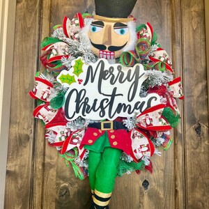 May include: A Christmas wreath featuring a nutcracker design. The wreath has a black top hat with gold trim, a tan face, and a black mustache. The wreath is decorated with red, green, and white ribbons and the words "Merry Christmas".